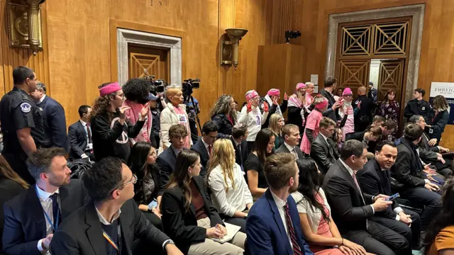 A group of people wearing pink stand at the back of the committee room, which is filled with people seated and waiting for the hearing to begin. The protesters are wearing pink and some have their hands raised