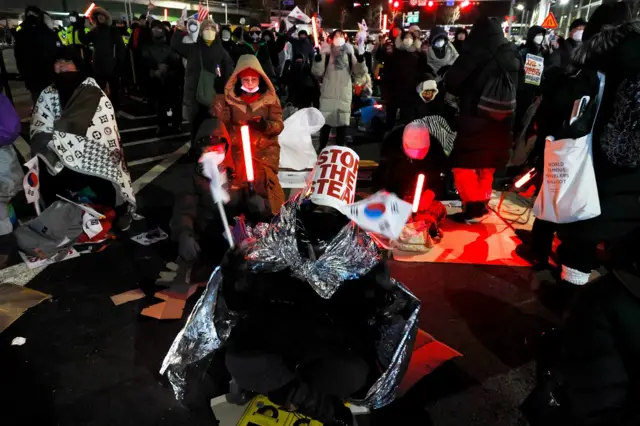 A Yoon supporter sitting on a ground, wrapped in a thermal blanket and waving a South Korean flag, with a "Stop the Steal" sign wrapped around their head.