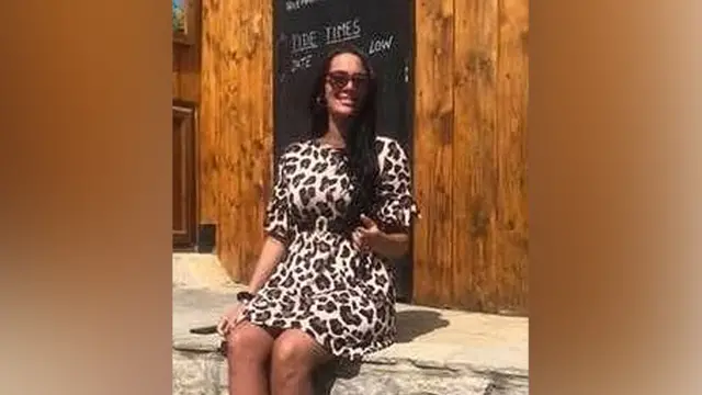 Kiena Dawes smiling with long black hair wearing sunglasses and a zebra-print dress sitting on a stone wall outside a cafell outsi