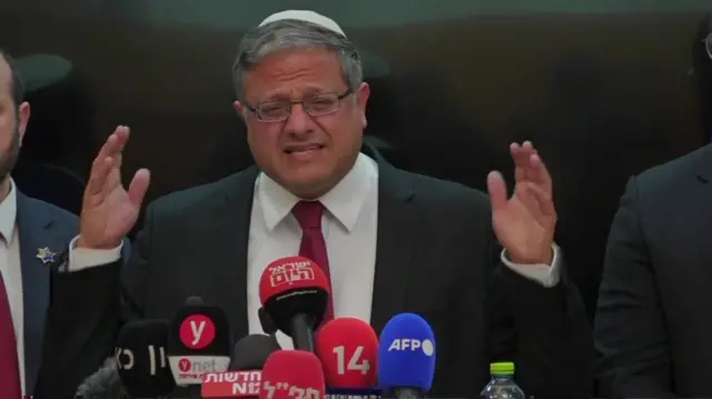 Ben-Gvir wearing a black suit and red tie, speaking at a news conference with his arms raised