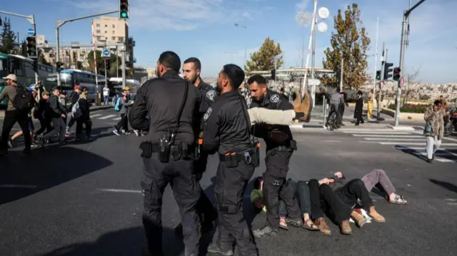 Four male law enforcement officers carry a person whose legs and feet can be seen. Other protestors lay on the ground to the right.