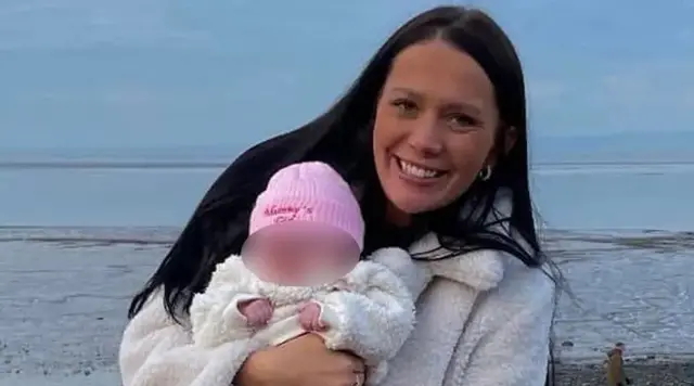 Kiena Dawes with long black hair smiling wearing a cream furry coat holding her baby dressed in a pink woolly hat, white furry coat and pink leggings and pink boots on the promenade with the sea in the background