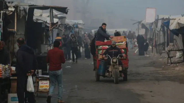 A three-wheeled motorbike is driven along a road lined with tents. The vehicle has crates on the back, which a man is sitting on.