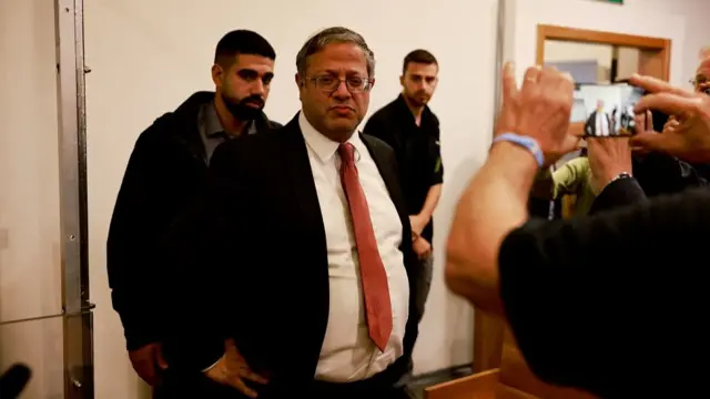 Itamar Ben Gvir wearing a suit and standing with his hands on his hips in front of photographers