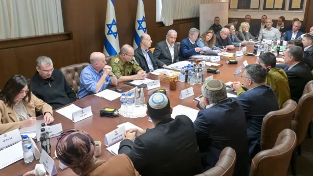 Men and women, some dressed in suits and others wearing army uniform, sit round a table with two Israeli flags in the background