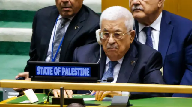 Mahmoud Abbas sitting in a chair at a green-topped desk while attending the UN general assembly. He's wearing a dark suit and blue tie, two men also in dark suits are sitting behind him