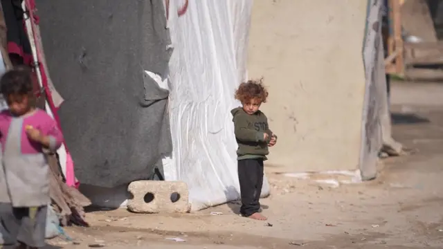 A very young Palestinian child stands barefoot outside a tent in Deir al-Balah