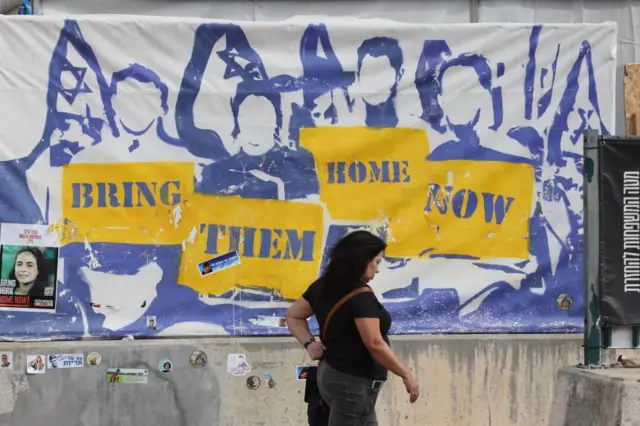 A woman walks past a poster on a wall saying Bring Them Home Now