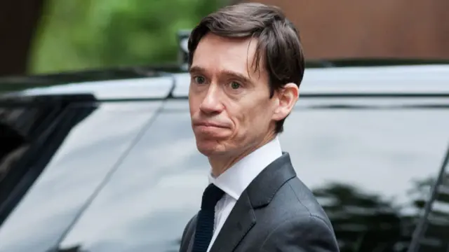 Rory Stewart stands in front of a black car with tinted windows. He is wearing a grey suit with a black tie and has brown hair and blue eyes.