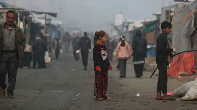 A young Palestinian girl is pictured standing in the middle of a street on a misty morning. Other people walk by and there are tents lining the street