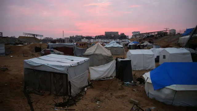 A camp in Gaza City, with tents of different sizes. The sun is setting, turning the sky pink. In the distance, a damaged building is visible.