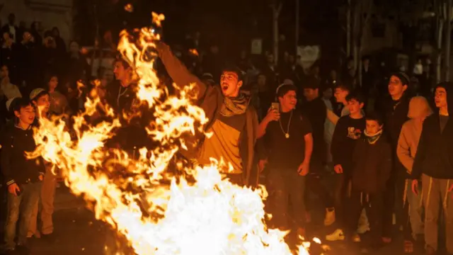 A crowd stand in front of a fire. One man stands at the front, holding his right arm up in the air as people watch on.