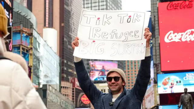 A man in a white beanie and sunglasses hold up a sign saying "TikTok refugee" while he smiles to a camera