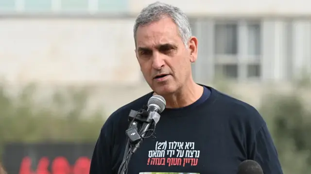 A man wears a black t-shirt with the face of a man being held hostage. The t-shirt has Hebrew words on it and he stands at a podium