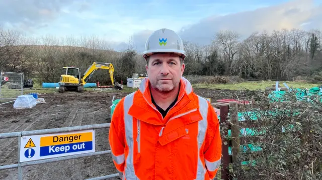 A man in an orange jacket and white hard hat is looking at the camera. Behind him is a sign that says "Danger Keep Out" and a yellow digger