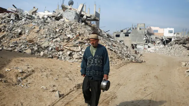 Palestinian man carrying a pot and wearing a blue patterned cardigan walks past the rubble of buildings destroyed in previous Israeli strikes, ahead of a ceasefire set to take effect on Sunday, in Khan Younis in the southern Gaza Strip