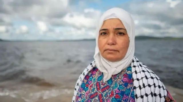 Majida Al Askri looking at camera, with a Palestinian keffiyeh draped over her shoulders, with the ocean in the background