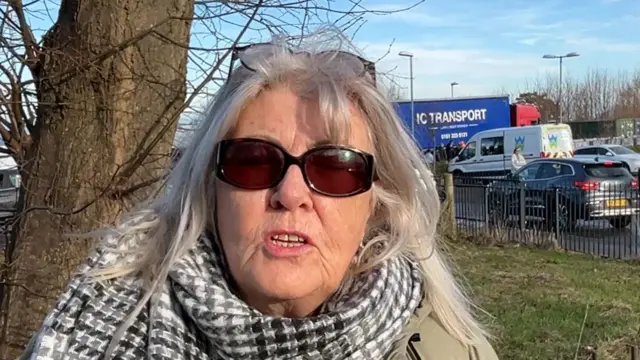 A woman with grey hair, wearing sunglasses and a black and white scarf is looking at the camera