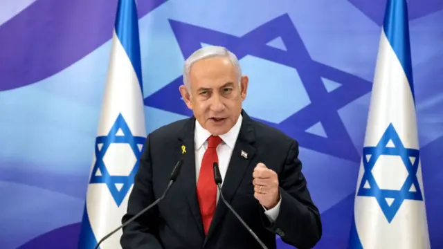 Netanyahu standing on stage delivering speech in black suit, his left arm extended forward with his hand in a fist, two Israeli flags at his side