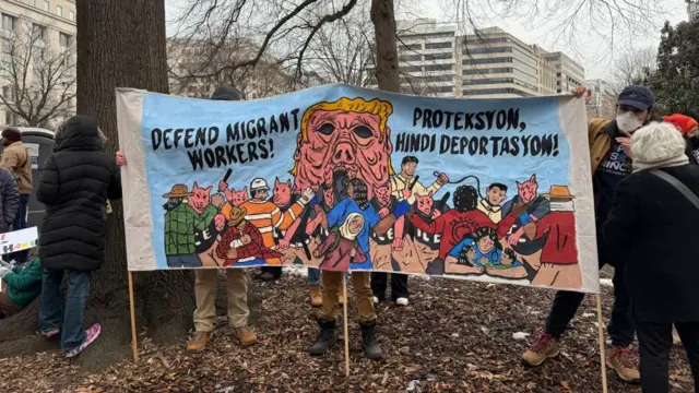 A group of people hold up a sign that says: Defend migrant workers. The image shows a melting Donald Trump face with people wearing pig masks confronting a group of migrant workers.