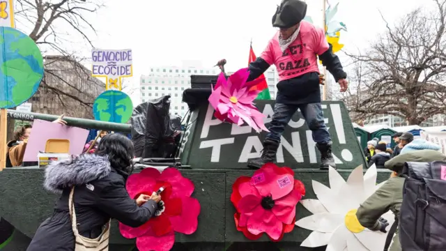 Person standing on top of a replica army tank which reads 'peace tank'