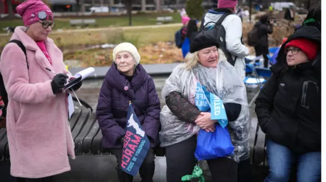 Older looking women sit on a park bench and hold signs and wear pink hats as they appear to be waiting for the march to get under way