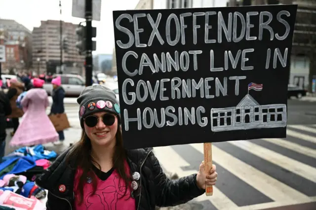 Woman in sunglasses holds a sign that reads "Sex Offenders Cannot Live in Government Housing"