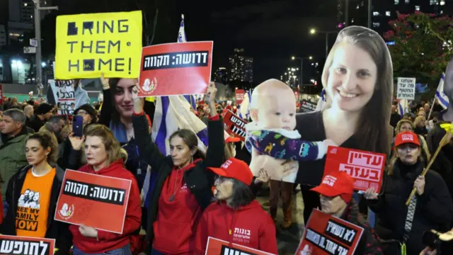 Anti-government protesters wearing red hoodies and shirts with pictures of the Bibas family march in Tel Aviv. One man in a black hat to the left of the frame holds up a yellow placard reading Bring them Home. At the forefront of the picture, to the right, is a printed cardboard version of a photo of Shiri Bibas holding her son Kfir