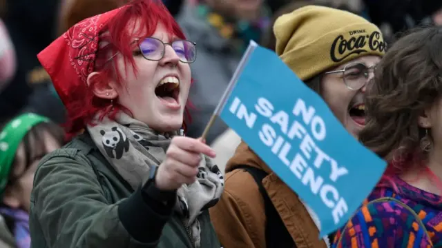 A woman with pink hair holds up a sign that reads: No safety in silence.