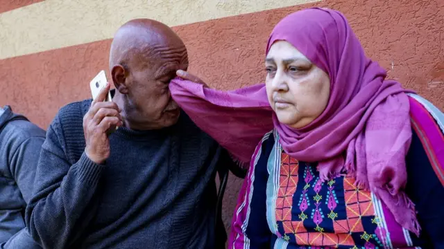 A man in dark grey shirt and matching vest holds a phone to his ear while wiping an eye with the corner of his wife's purple headscarf. They're sitting down in front of a terracotta-coloured wall with a cream white stripe running across