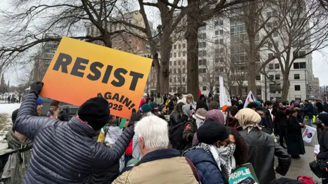 A crowd of people is shown on the streets of downtown Washington, with a person holding up a sign that reads: Resist again 2025