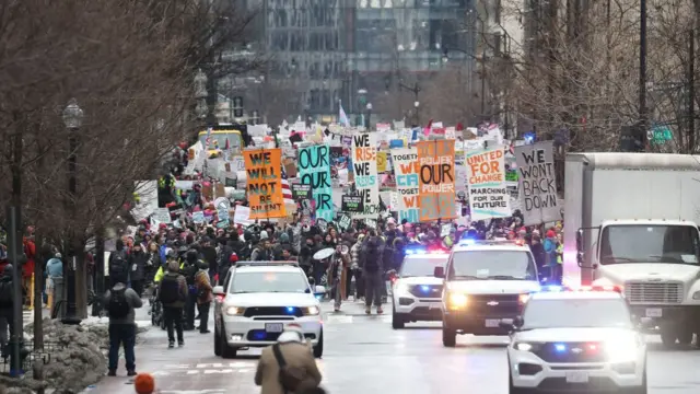 Police cars drive ahead of a large crowd marching down a road in DC