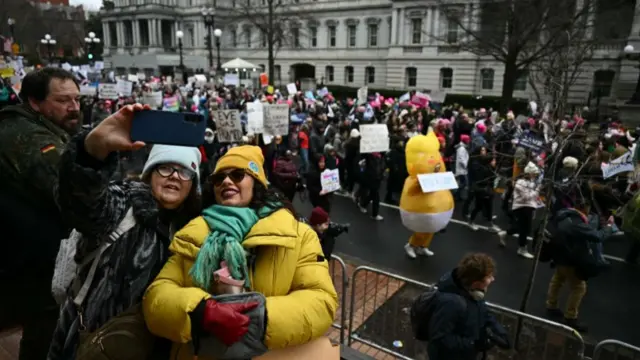 People pose for selfies in front of a large crowd of people marching in the People's March. They hold various signs expressing their support for various causes