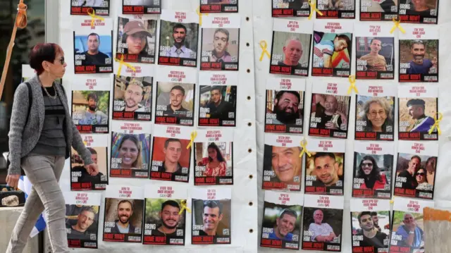 Woman in grey jacket, dark grey shirt and grey jeans walks along display of pictures of hostages still being held in Gaza haning on a white plastic tent outdoors