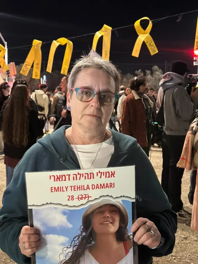 Emily Damari's mum holding a poster with a photo of her daughter Emily while at a march in southern Israel. Demonstrators can be seen milling around her, yellow ribbons are tied to a string hanging above her head