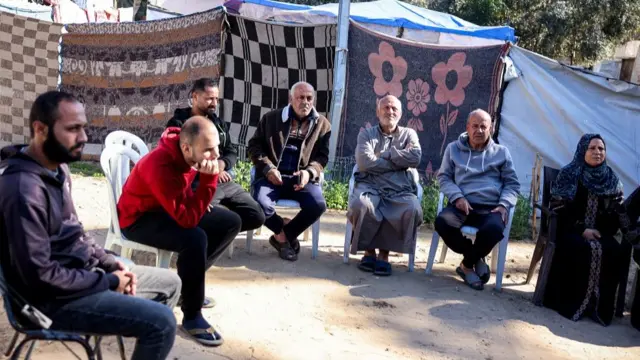 A group of people sit on plastic chairs in a semi circle -from left to right, three men and a woman in the far right of the image, sitting in front of makeshift tents