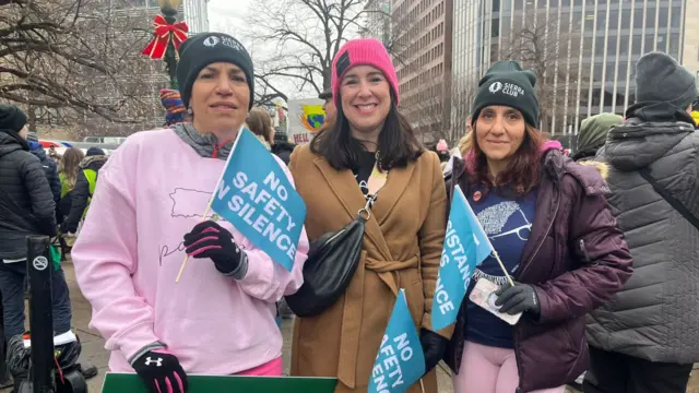 Three women standing next to each other, holding small blue flags that read: No safety no silence