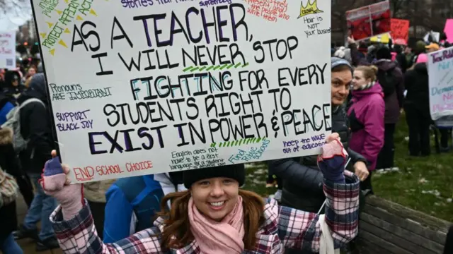 A woman holds up a sign that says: As a teacher, I will never stop fighting for every students right to exist in power & peace. There are a number of efforts on the sign that describe what the teacher is supporting, including pronouns, black joy, protecting affirmative action and representation within schools