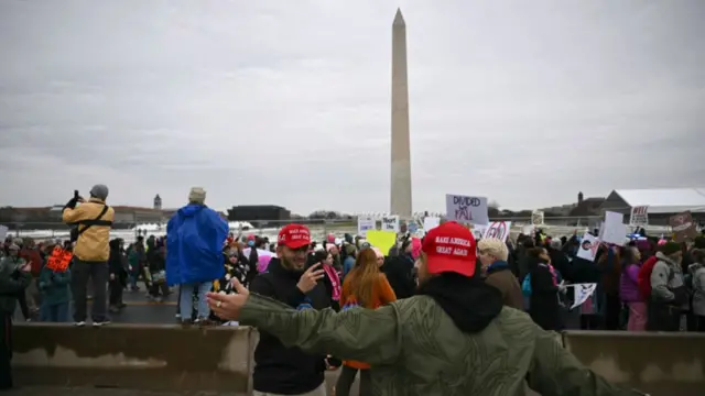 Two men pose in front of a group of protesters who are marching in DC against Donald Trump. The men are smiling and taking selfies as they wear Make America Great Again (Maga) hats.