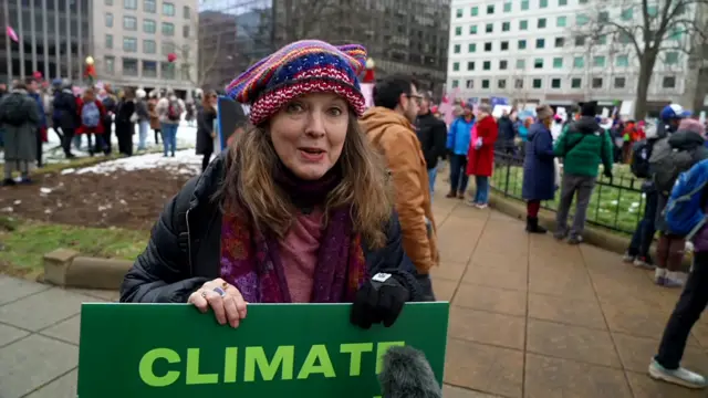 A woman stands in a crowd of people and holds up a green sign that says, Climate Action Now. She wears a knit hat that has cat ears on the side - referred to as "pussy hat"