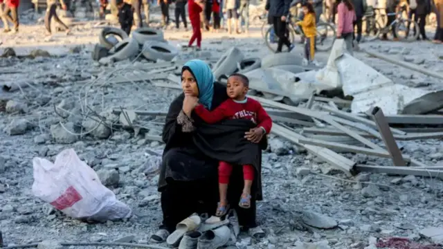 A woman and child sit in the middle of rubble in Gaza with a crowd of people in the background