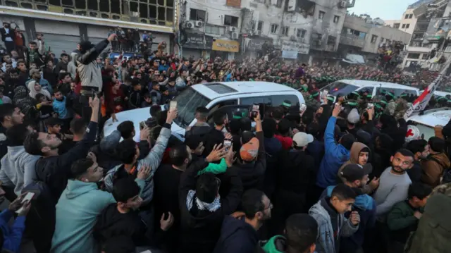 Large group of men crowd around several white Red Cross vans during hostage exchange in Gaza City. Several of the men are holding up phones as they film the scene, damaged homes and buildings are visible in the background