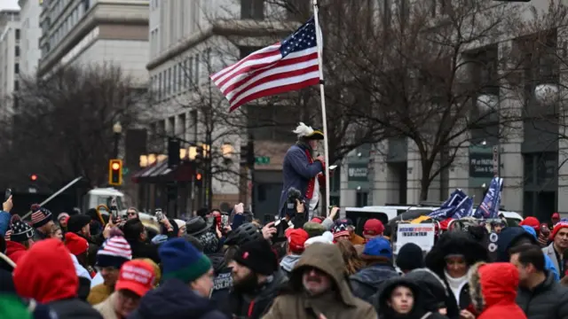 A man in a revolutionary era outfit with a US flags stands raised up above a crowd, many wearing red Maga hats