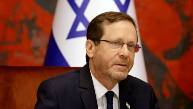 Israeli president Isaac Herzog sitting down in a black suit with matching tie and white shirt in front of Israeli flag and red background