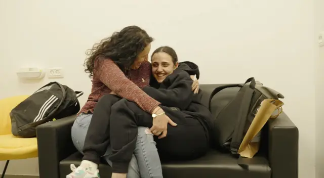 Romi Gonen, 24 hugs her mother while sitting on a black leather couch. She's in a black jumper and black joggers and is smiling