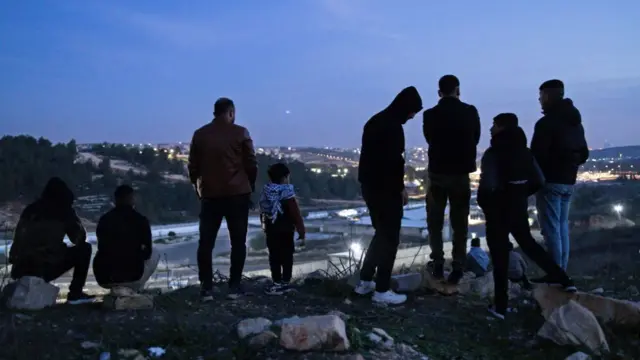 Palestinians on a hill overlooking the Ofer prison earlier today. They have their back to the camera, Ofer prison is visible at the bottom of the hill they're standing on, what appears to be a village illuminated by bright lights is visible in the background