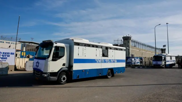 A white police mini-van with a blue stripe to the side and a Star of David in the front parked outside Ofer Prison. A similar van is parked behind it, the prison with two front turrets visible in the background