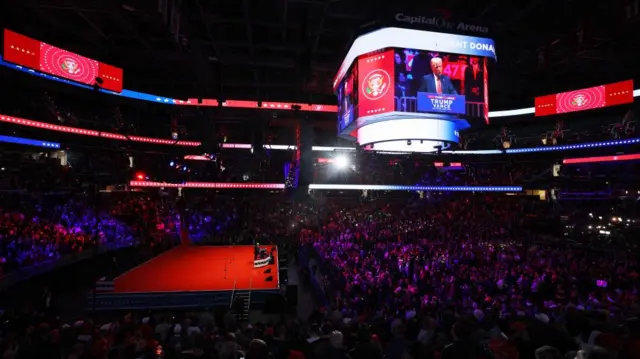 A wide shot showing the large venue and crowd, with Trump on stage, his face projected on the big screen high above the crowd