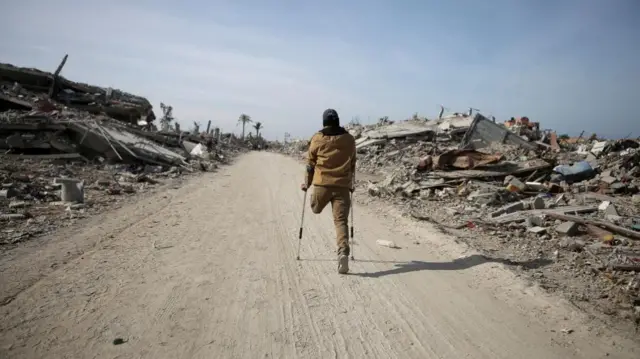 Displaced Palestinian man makes way past rubble in crutches