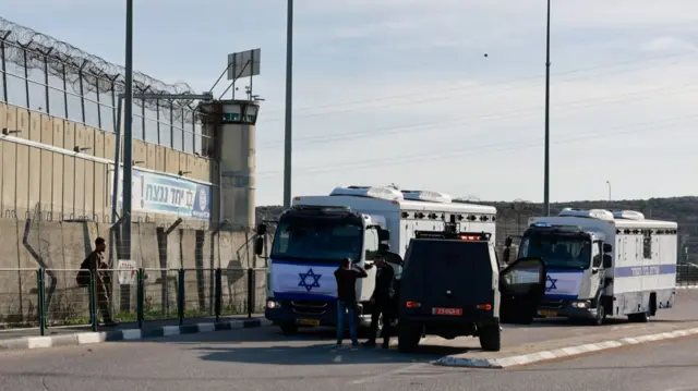 Israeli police cars wait to access Ofer Prison in the occupied West Bank. There's two larger white mini buses with Star of David emblazoned at the front are parked behind a black car with sirens on top, three men milling around